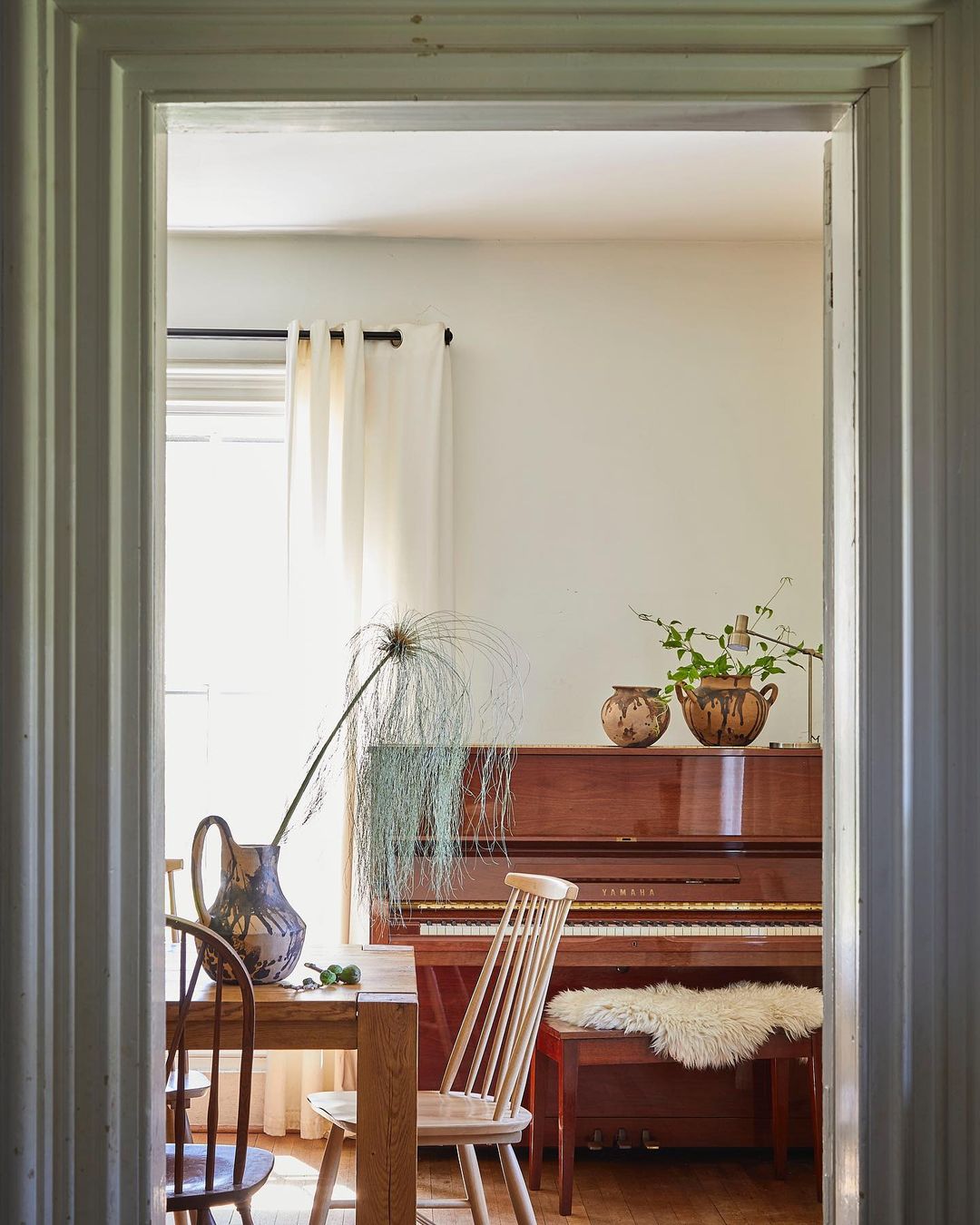 Interior view of a dining room with a table and chairs, a piano in the background, and natural light coming through a window.