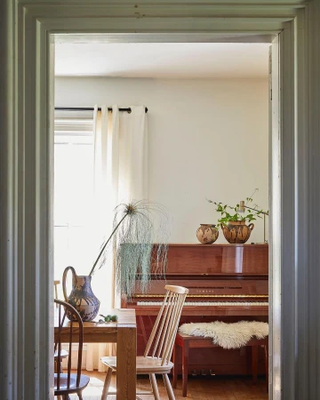 Interior view of a dining room with a table and chairs, a piano in the background, and natural light coming through a window.