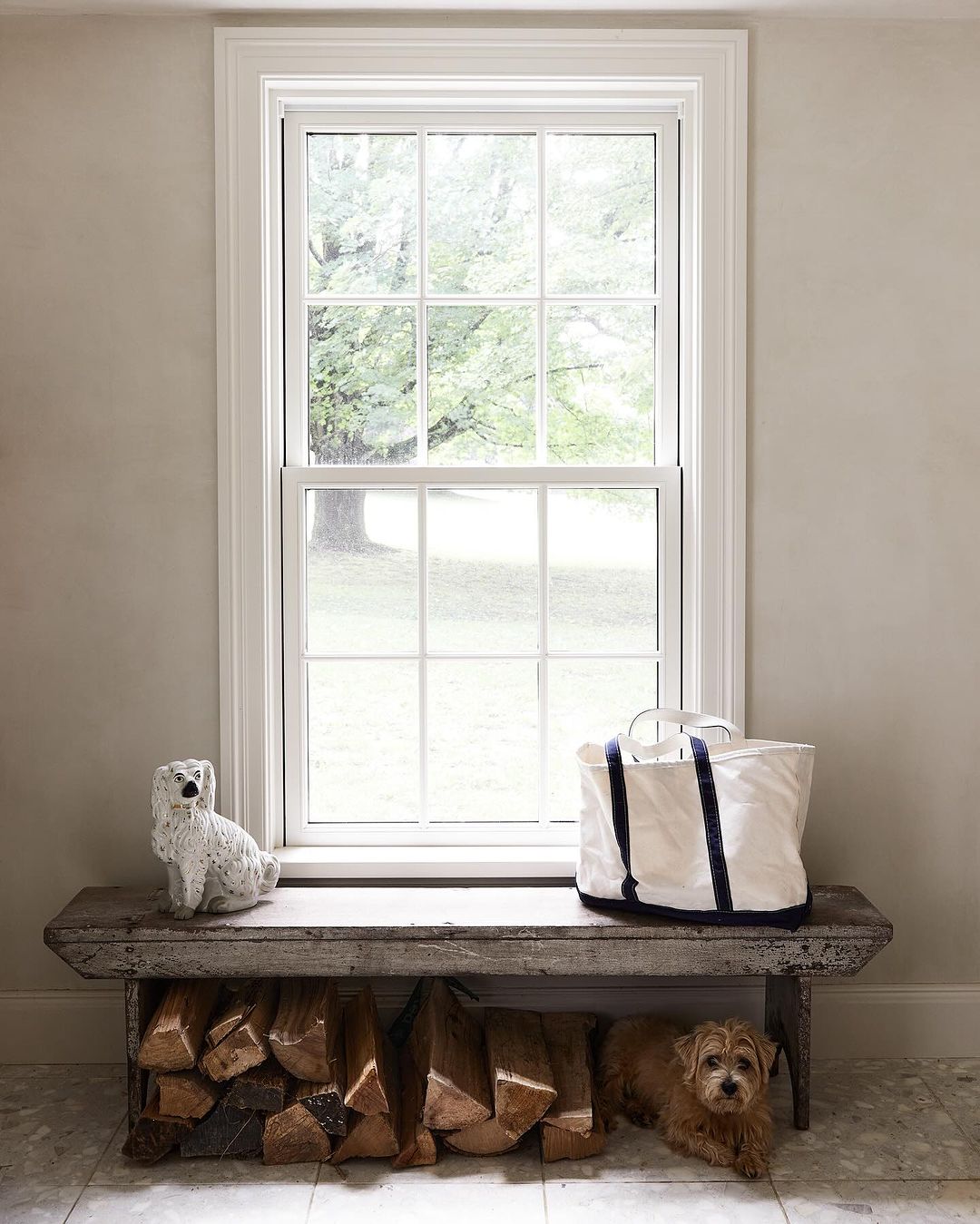 Indoor scene featuring a dog lying beneath a wooden bench with a tote bag, a dog figurine, and a window view of greenery.