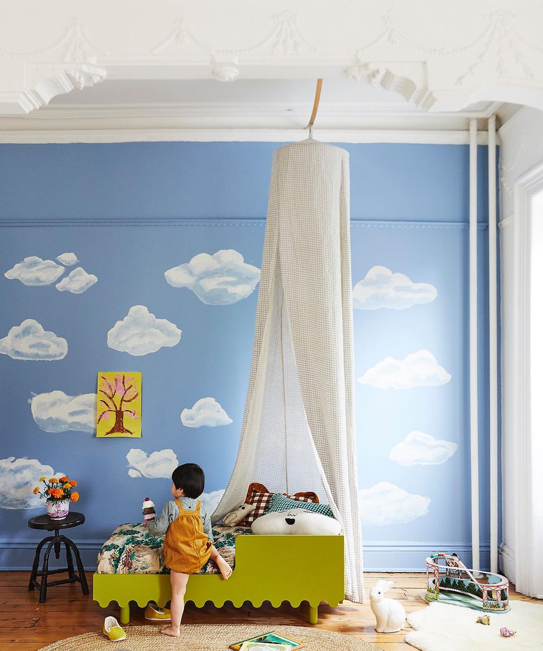 Child in a bright kids room with a lime green bed under a canopy, blue wall with painted clouds, and a braided jute rug.