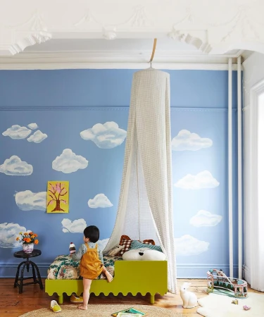 Child in a bright kids room with a lime green bed under a canopy, blue wall with painted clouds, and a braided jute rug.