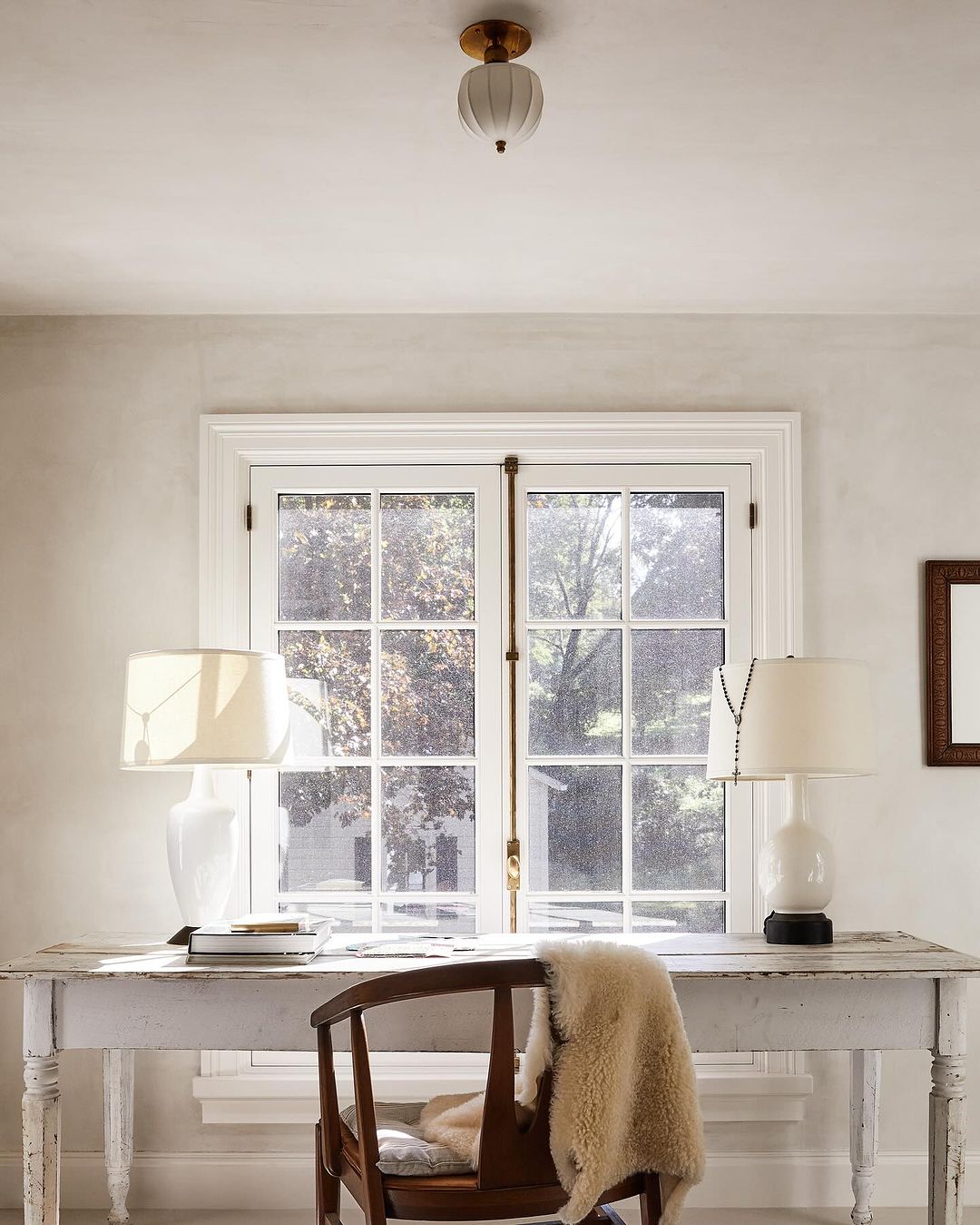 Interior photo of a white desk with two lamps, a chair with a sheepskin throw, and a window in the background.