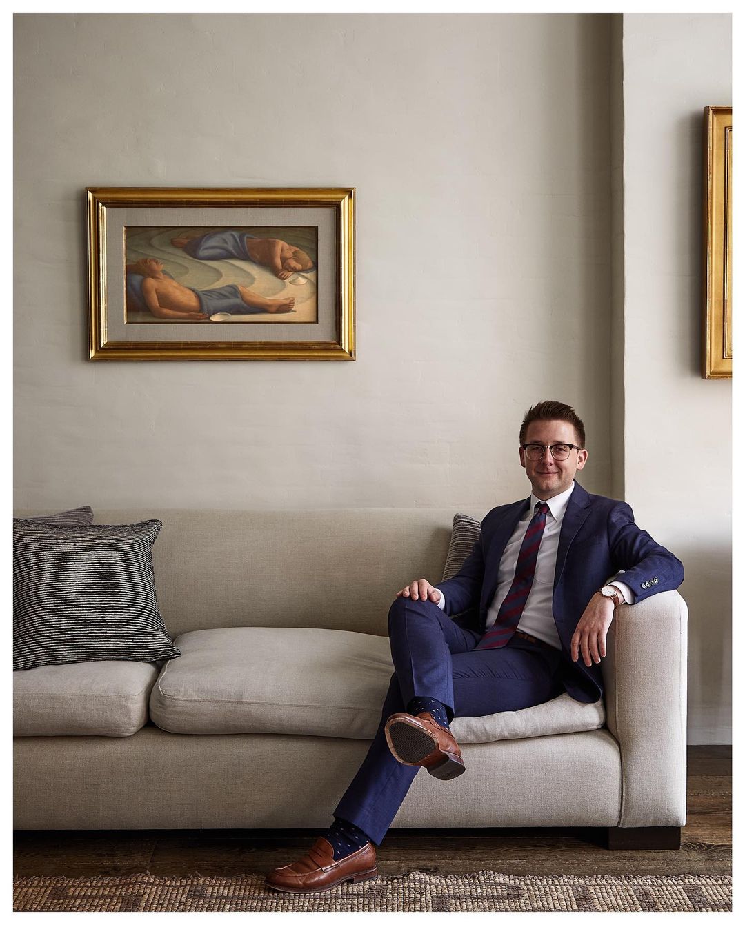 Adult man in a blue suit sitting on a cream sofa with an art painting in the background, wearing glasses and looking forward indoors.