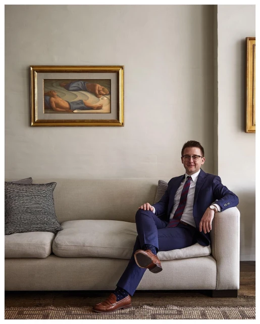 Adult man in a blue suit sitting on a cream sofa with an art painting in the background, wearing glasses and looking forward indoors.