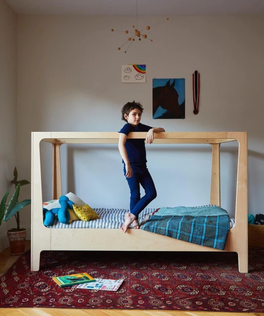 Young child stands on a modern wooden bed in a naturally lit room with colorful art, mobile, and a patterned rug on the wooden floor.