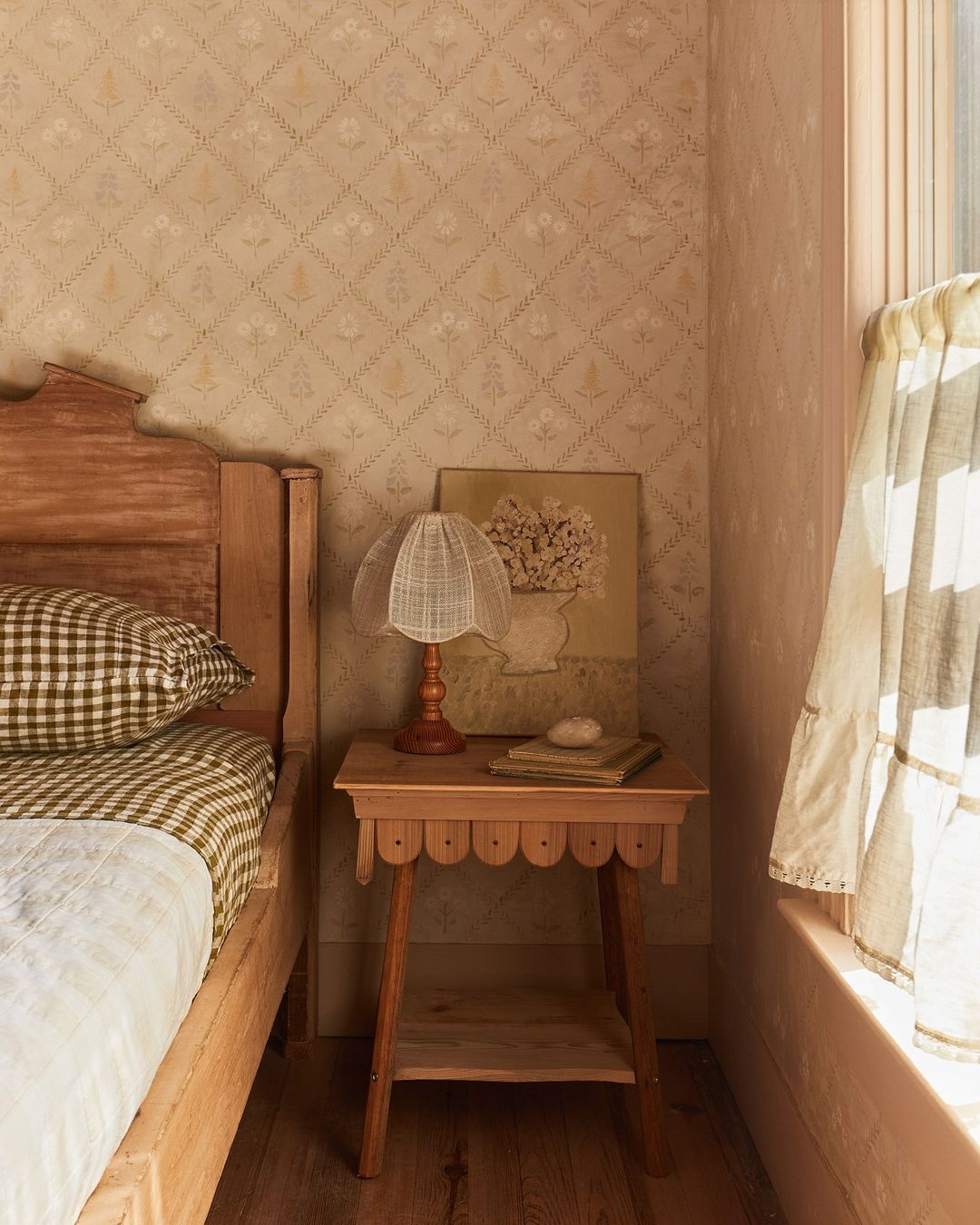 Rustic bedroom interior featuring a wooden bed with gingham bedding, a scalloped side table with a lamp, and patterned wallpaper.