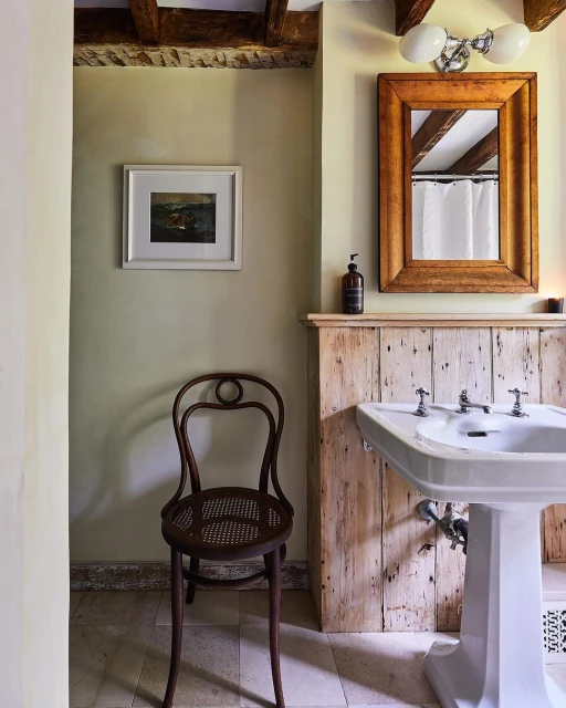 Interior bathroom features a vintage chair, white pedestal sink, aged wooden wall, framed art, light fixtures, and pale yellow walls.