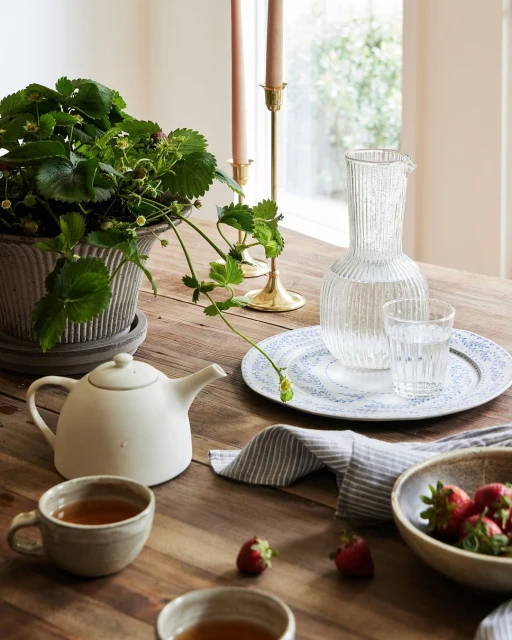 Still life of tea set with teapot, cups, clear pitcher, strawberries, and potted plant on a wooden table near a window.