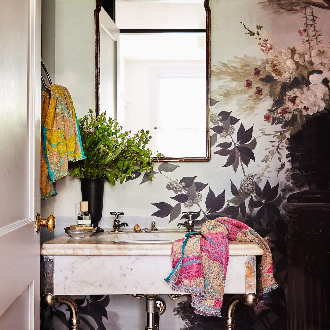 Bathroom interior featuring a marble sink, mirror, floral wallpaper, and colorful towels creating a bright and decorative space.