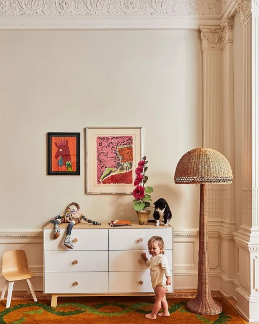 Toddler stands near a white dresser with colorful art, a plant, and a cat in an interior with a wicker floor lamp.