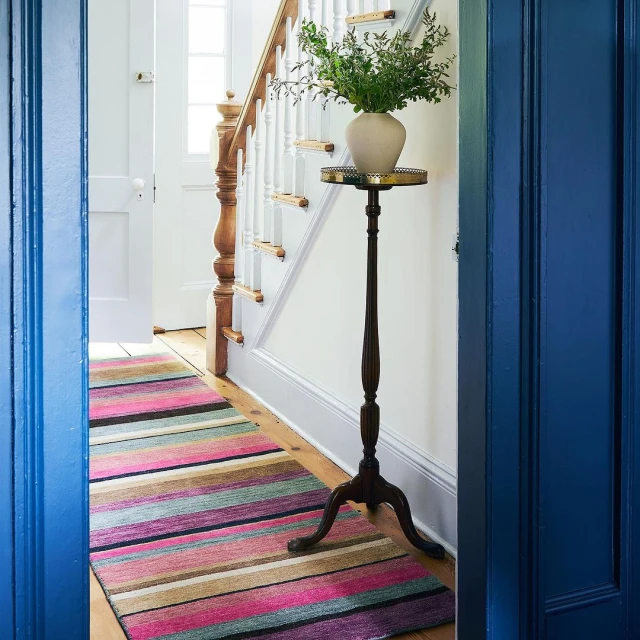 Indoor interior shot of a striped rug on a wood floor, a plant stand with a vase, stairs with white banisters, and a blue door frame.
