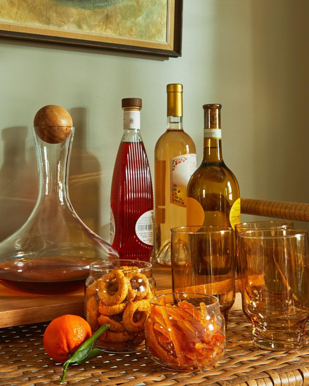 Still life arrangement features a decanter, bottles of wine, glass jars with snacks, and drinks in glasses on a rattan table.