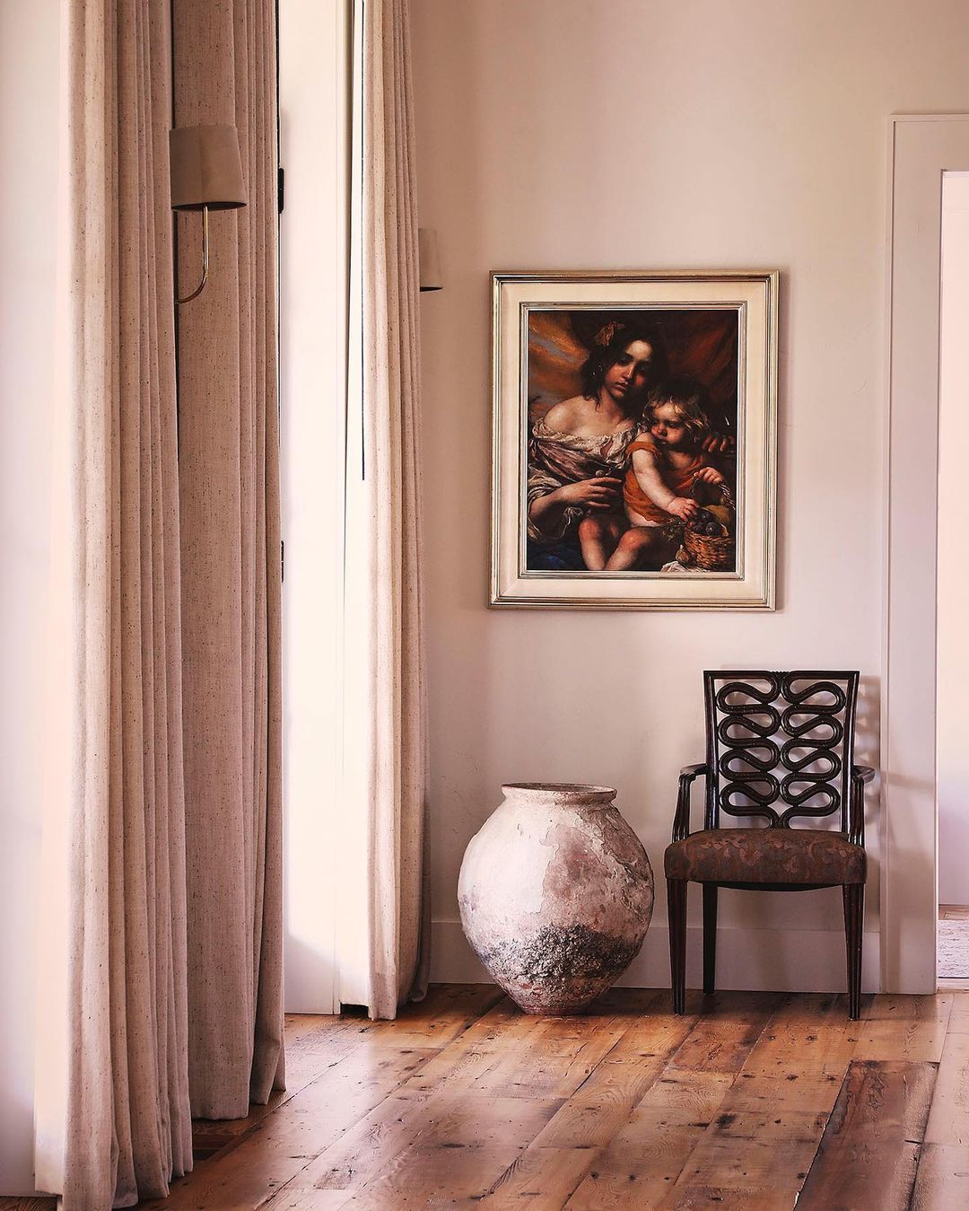 Interior view of a room with a framed painting of a Madonna and child, a chair, a large vase, and beige curtains.