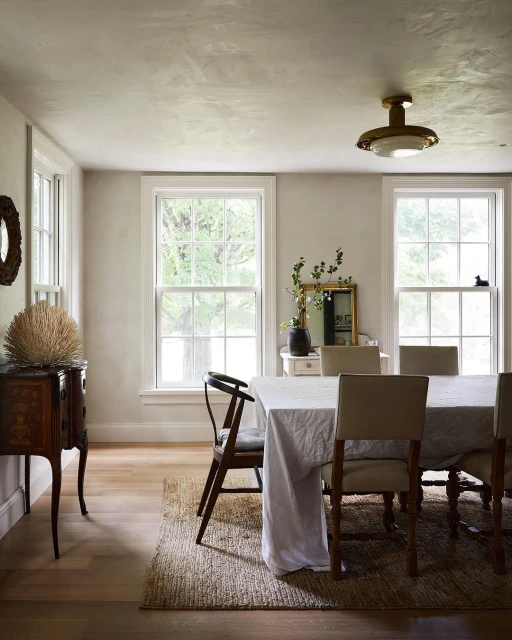 Dining room interior with a dining table, chairs, and a side table with a decorative object, illuminated by natural light.