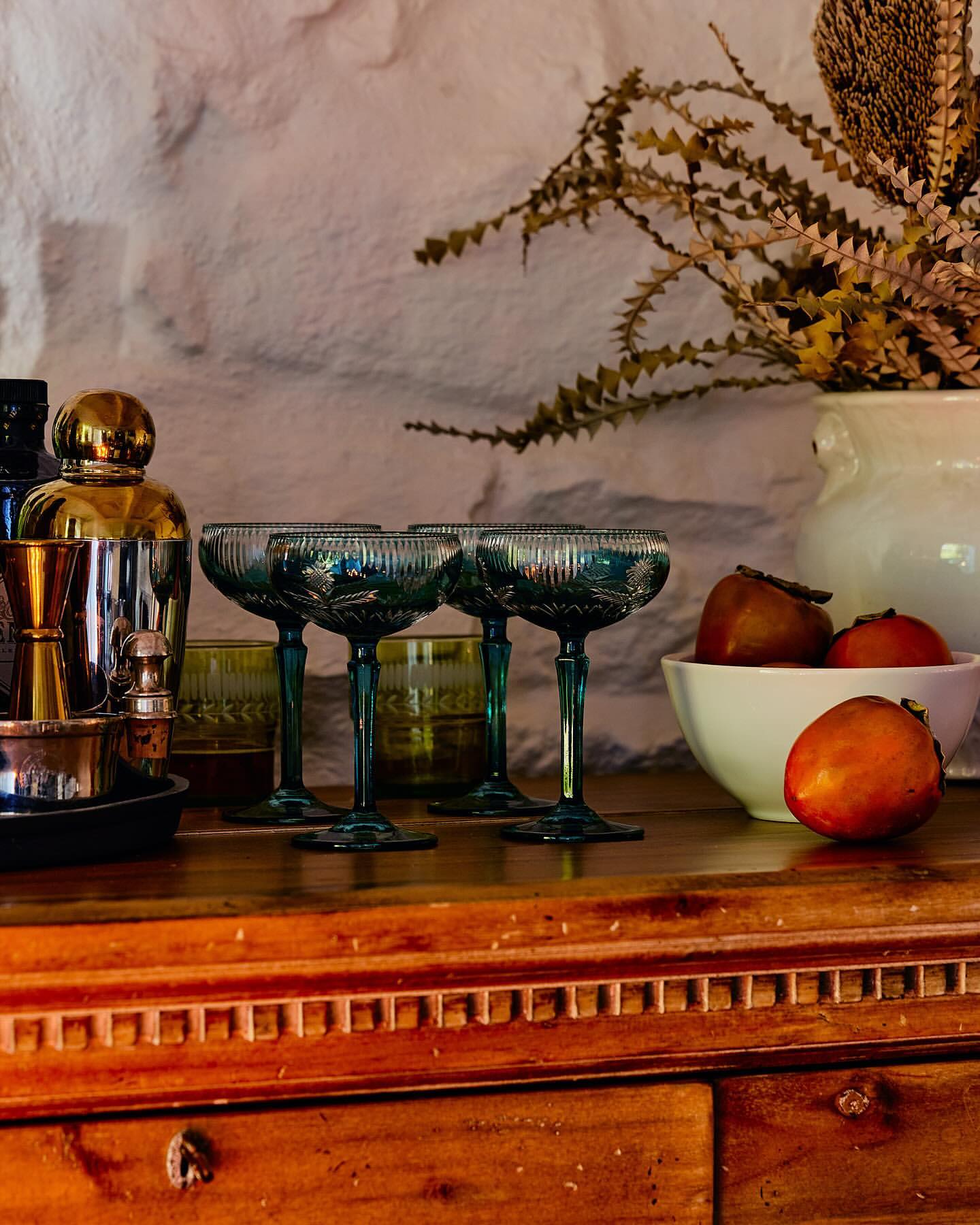 Arrangement with four stemmed coupe glasses, cocktail accessories, fruit bowl, decorative vase, and weathered wooden cabinet.