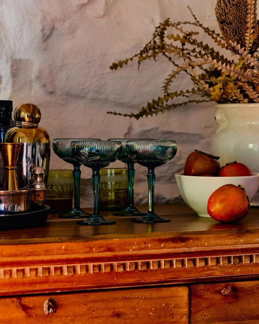 Arrangement with four stemmed coupe glasses, cocktail accessories, fruit bowl, decorative vase, and weathered wooden cabinet.