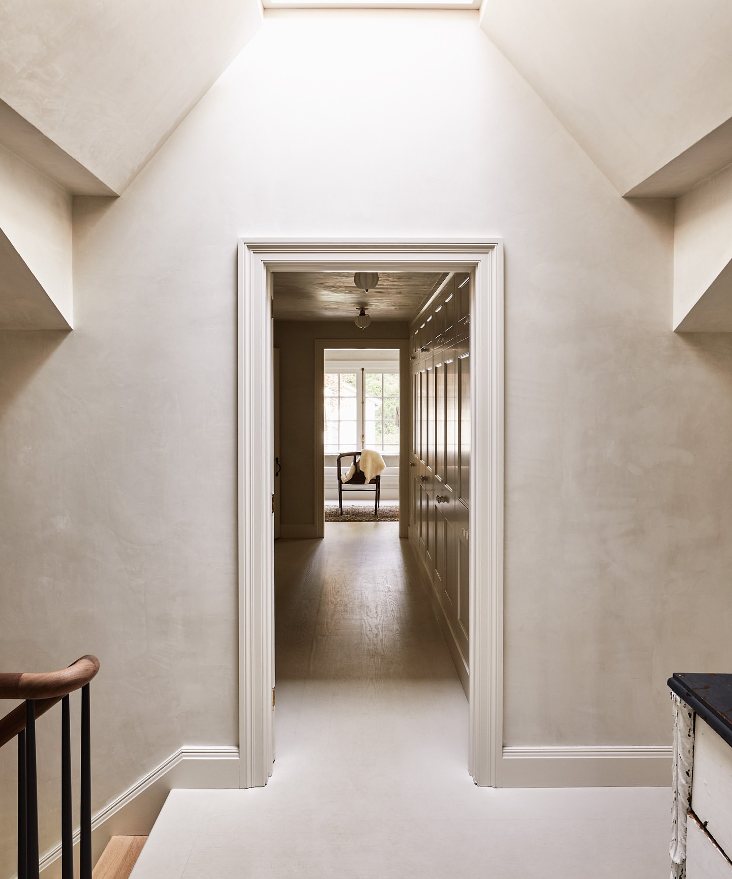 Interior view of a hallway with off-white walls, skylight, and open doorway leading to a room with a chair near a window.