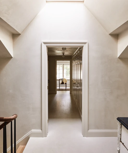 Interior view of a hallway with off-white walls, skylight, and open doorway leading to a room with a chair near a window.