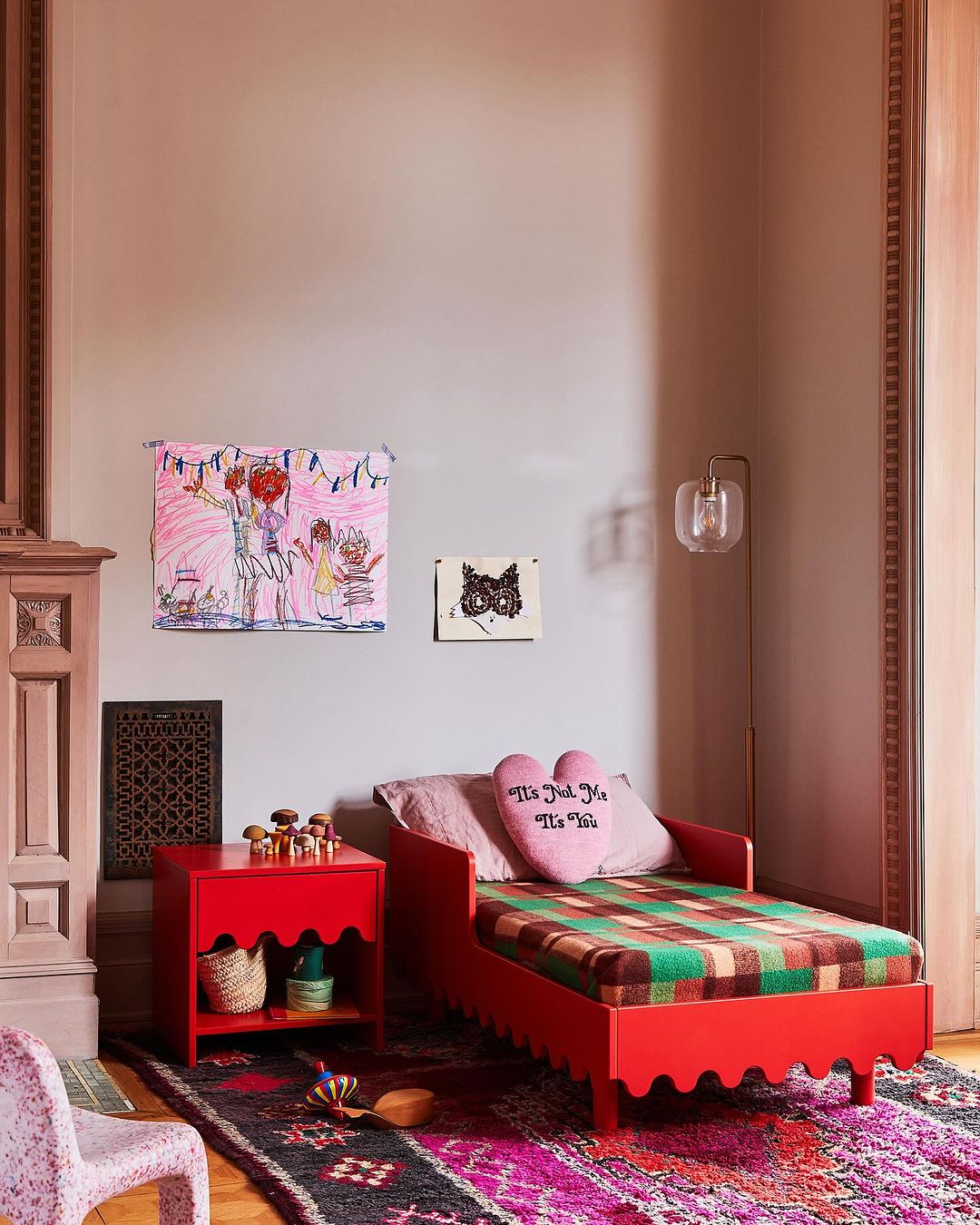 Red daybed with patterned blanket and heart-shaped pillow, alongside a red bedside table with decor, a patterned rug, and artwork.