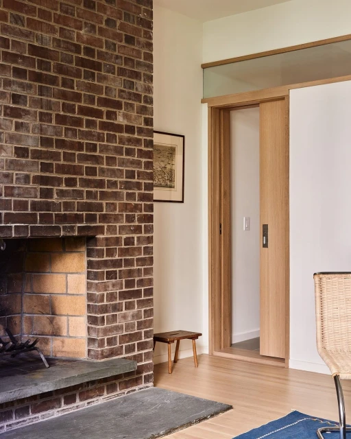 Interior view of a living room with a brick fireplace, wooden sliding door, wooden stool, light-colored wood floor and neutral-colored walls.