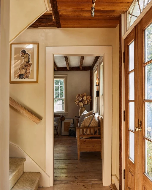 A naturally-lit interior hallway features a wooden staircase, doorway, framed art, and view of a cozy living room with wood furnishings.