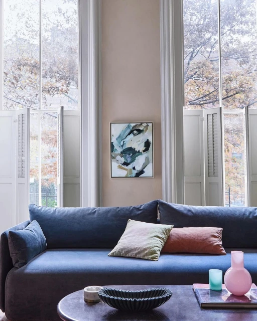 Living room interior featuring a blue sofa, decorative pillows, modern art, and natural light from large windows with white shutters.