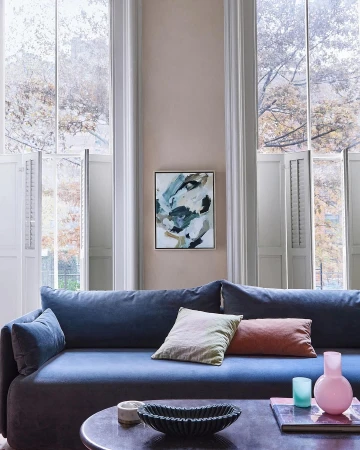 Living room interior featuring a blue sofa, decorative pillows, modern art, and natural light from large windows with white shutters.