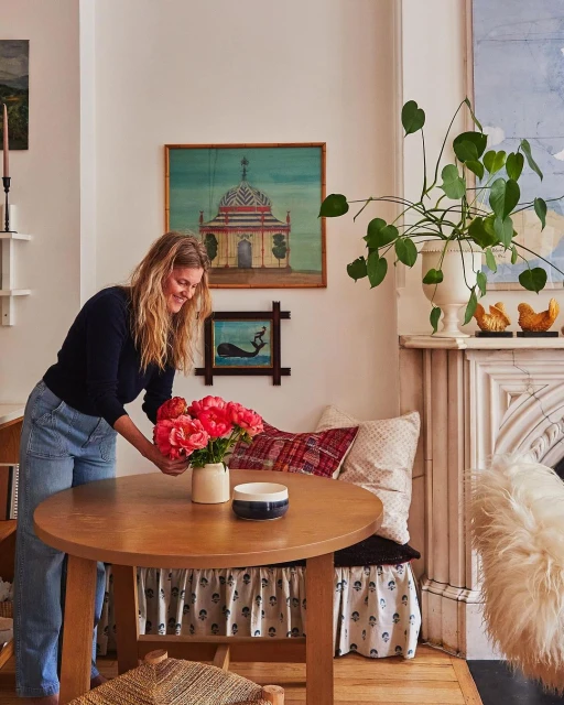 Woman arranging pink flowers on a round wood table in a home interior with art, potted plants, and natural light.