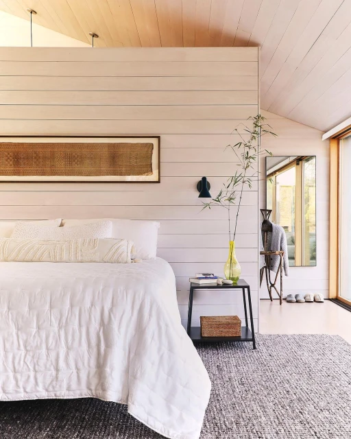 Bedroom interior featuring a white bed with pillows, a side table with a lamp, a rug, and minimalist decor in a modern home.