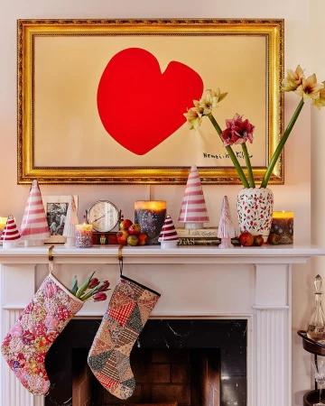 Festive mantelpiece with a large red heart artwork, Christmas stockings, decorative Christmas trees, candles, and flowers.
