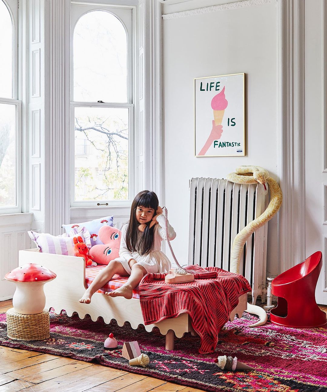 Young girl sits on a child's bed in a brightly lit room, talking on a pink rotary phone next to a radiator and colorful artwork.