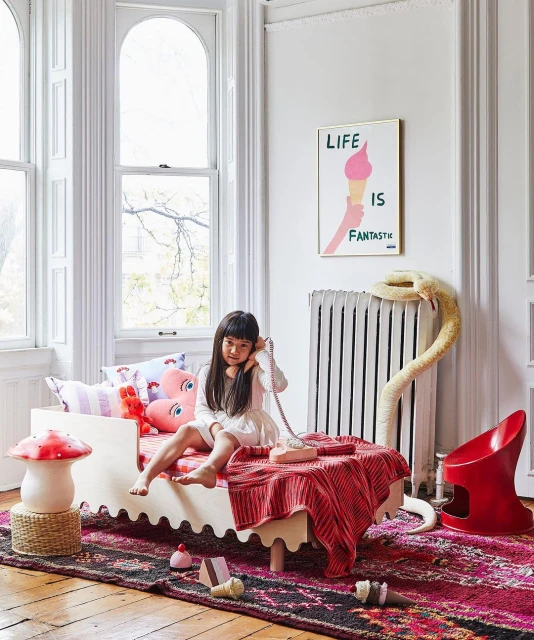 Young girl sits on a child's bed in a brightly lit room, talking on a pink rotary phone next to a radiator and colorful artwork.