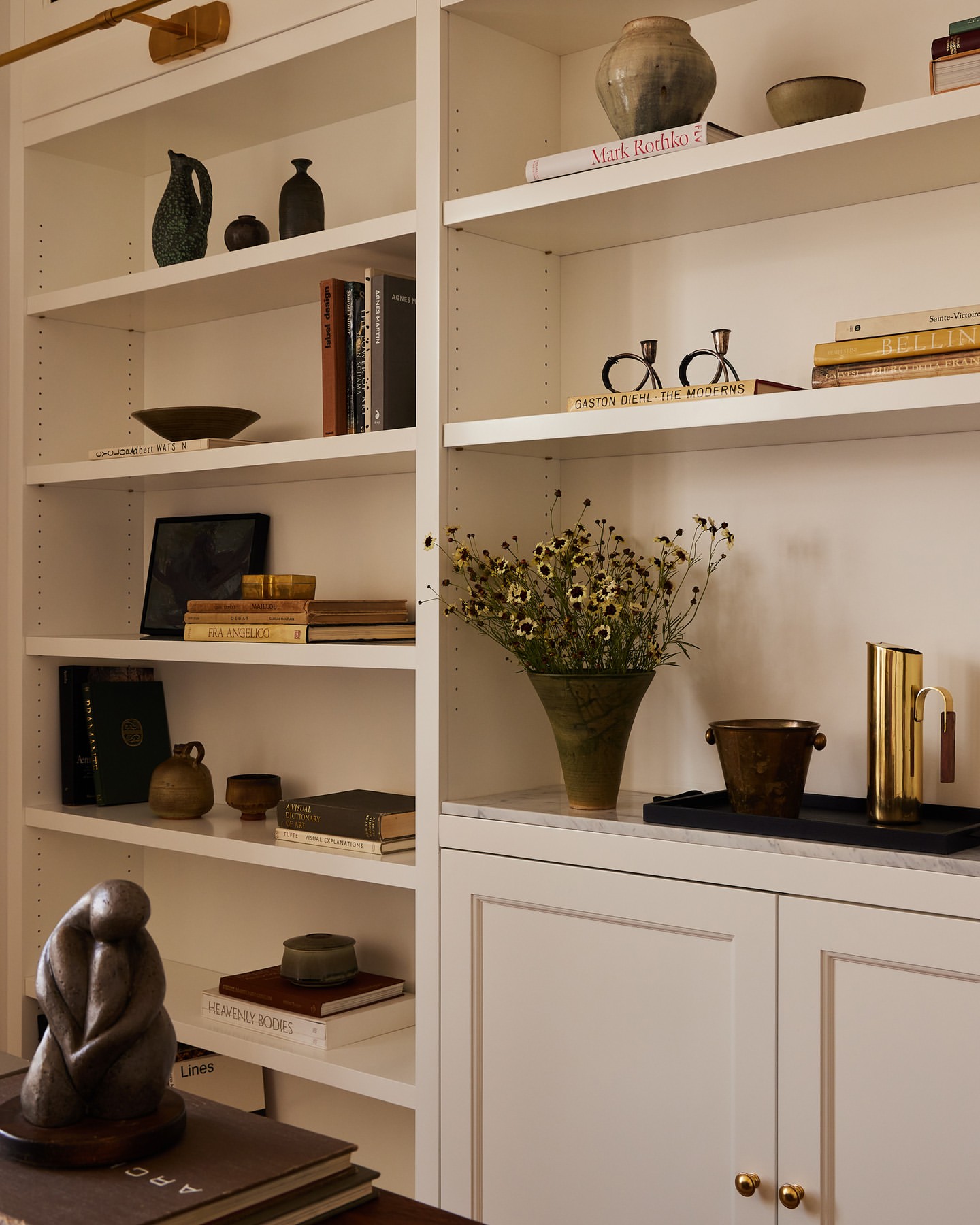 Living room interior with built-in white shelves filled with vases, pottery, books, and a bronze sculpture on a dark wood table.