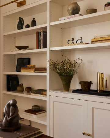 Living room interior with built-in white shelves filled with vases, pottery, books, and a bronze sculpture on a dark wood table.