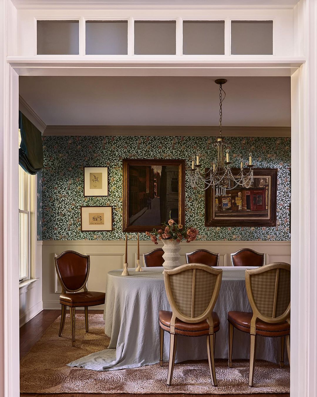 Elegant dining room interior with a round table, upholstered chairs, floral wallpaper, framed artwork, and a decorative chandelier.