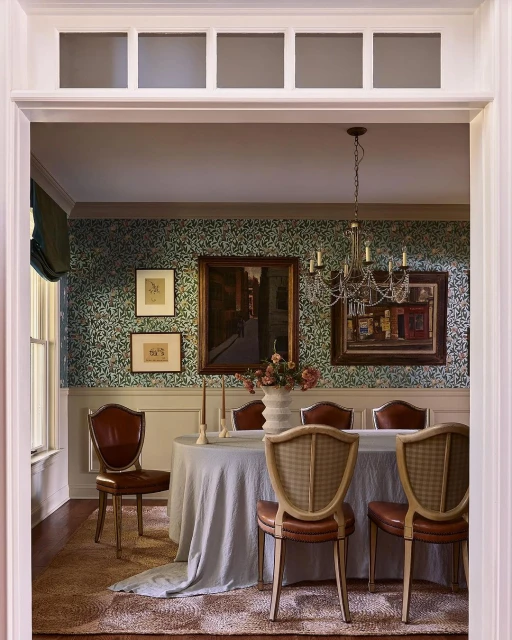 Elegant dining room interior with a round table, upholstered chairs, floral wallpaper, framed artwork, and a decorative chandelier.