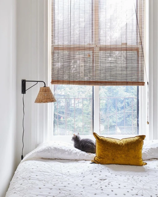 Cozy bedroom interior featuring a grey cat sitting on a white patterned bed, a yellow pillow, a window with blinds, and a wall sconce.