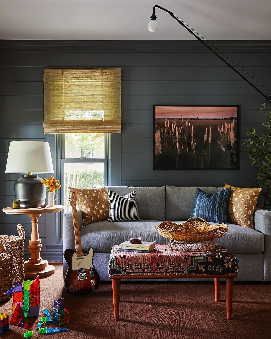 Living room interior with a grey couch, patterned pillows, and a vintage-style ottoman, featuring a guitar and colorful building blocks.