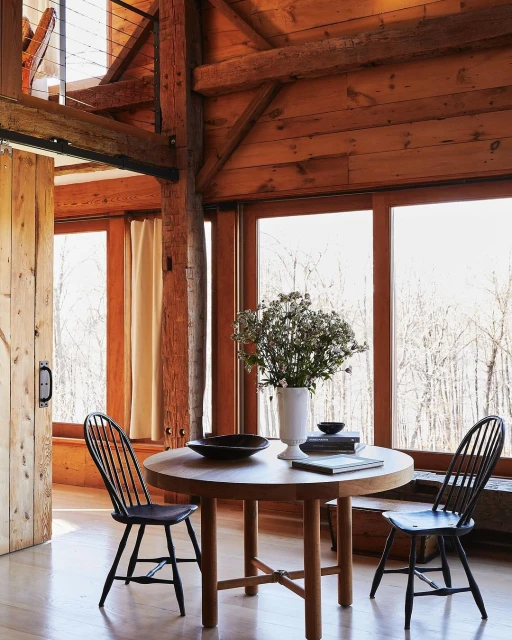 Interior of a wooden cabin features a round table with black chairs, a vase of flowers, and an open door, showcasing a rustic aesthetic.