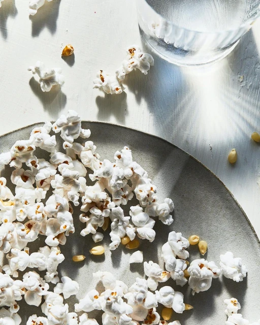 Overhead shot of popcorn scattered on a grey plate and white surface with a clear glass of water, creating a bright and airy food still life.