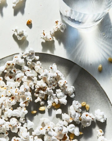 Overhead shot of popcorn scattered on a grey plate and white surface with a clear glass of water, creating a bright and airy food still life.