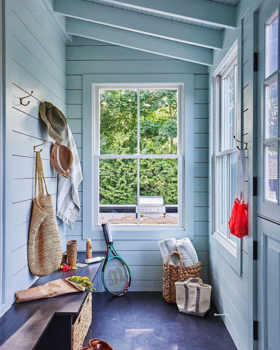 Cozy sunroom with blue wood paneled walls, large windows, beach-themed decor, straw bags, tennis racket, towels, and natural light.