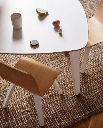 Wooden chairs surround a white table with toy food and a white cup on a woven brown rug atop wood flooring.
