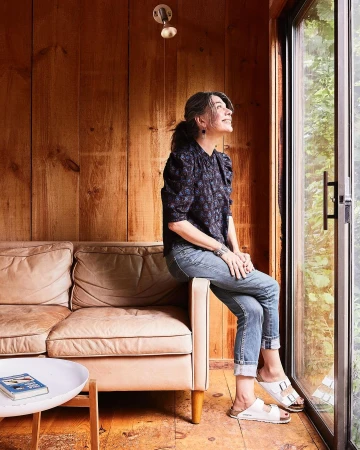 Adult woman in a patterned blouse sits on a peach-colored sofa near a window inside a wood-paneled home, looking up toward light.