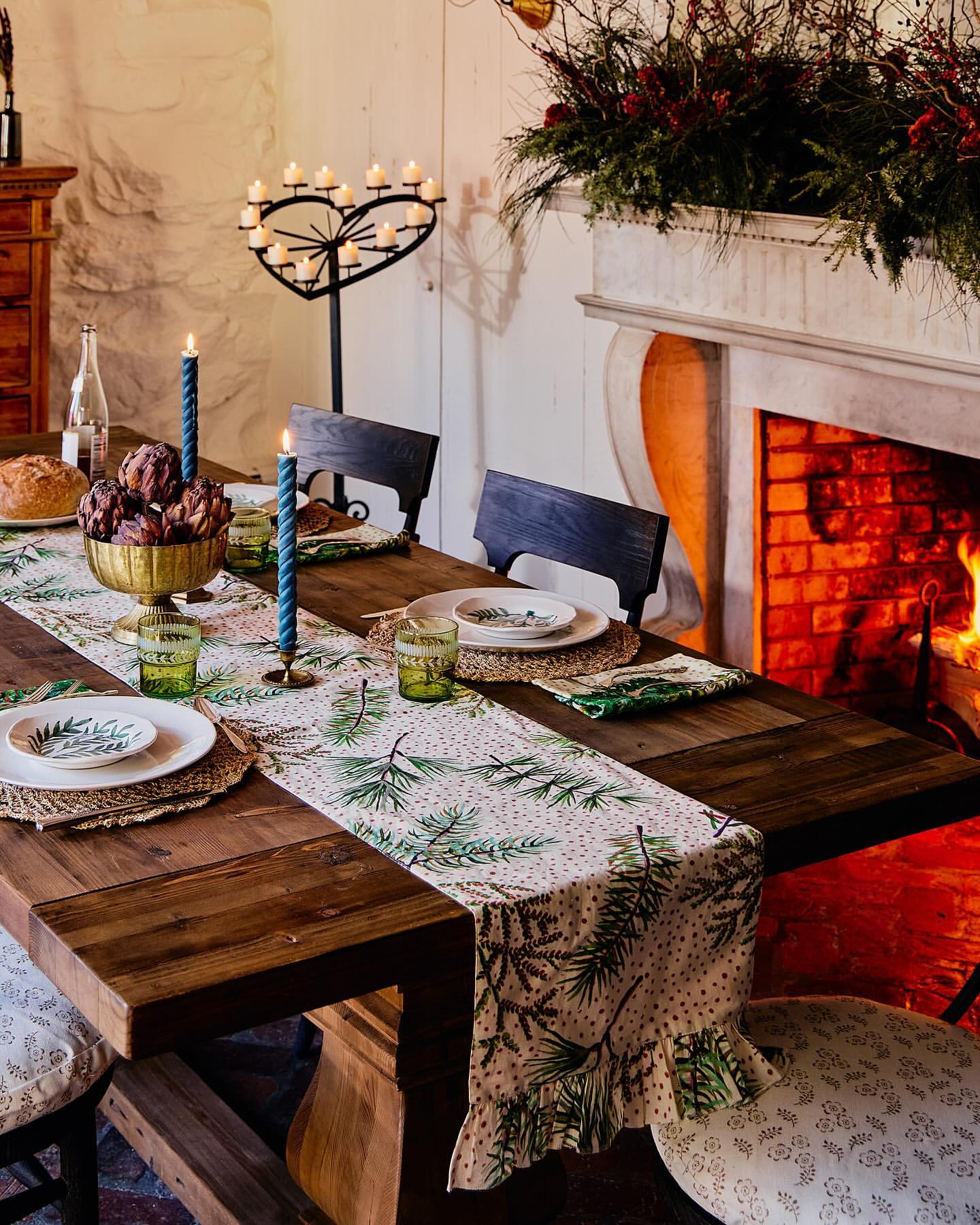 Holiday table setting with gold bowl of artichokes, green glassware, candles, white plates, and fireplace, creating a warm ambiance.