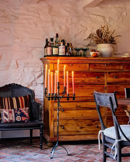 Candelabra with lit peach candles stands on a brick floor in a warmly lit interior featuring a wood dresser and vintage chair.