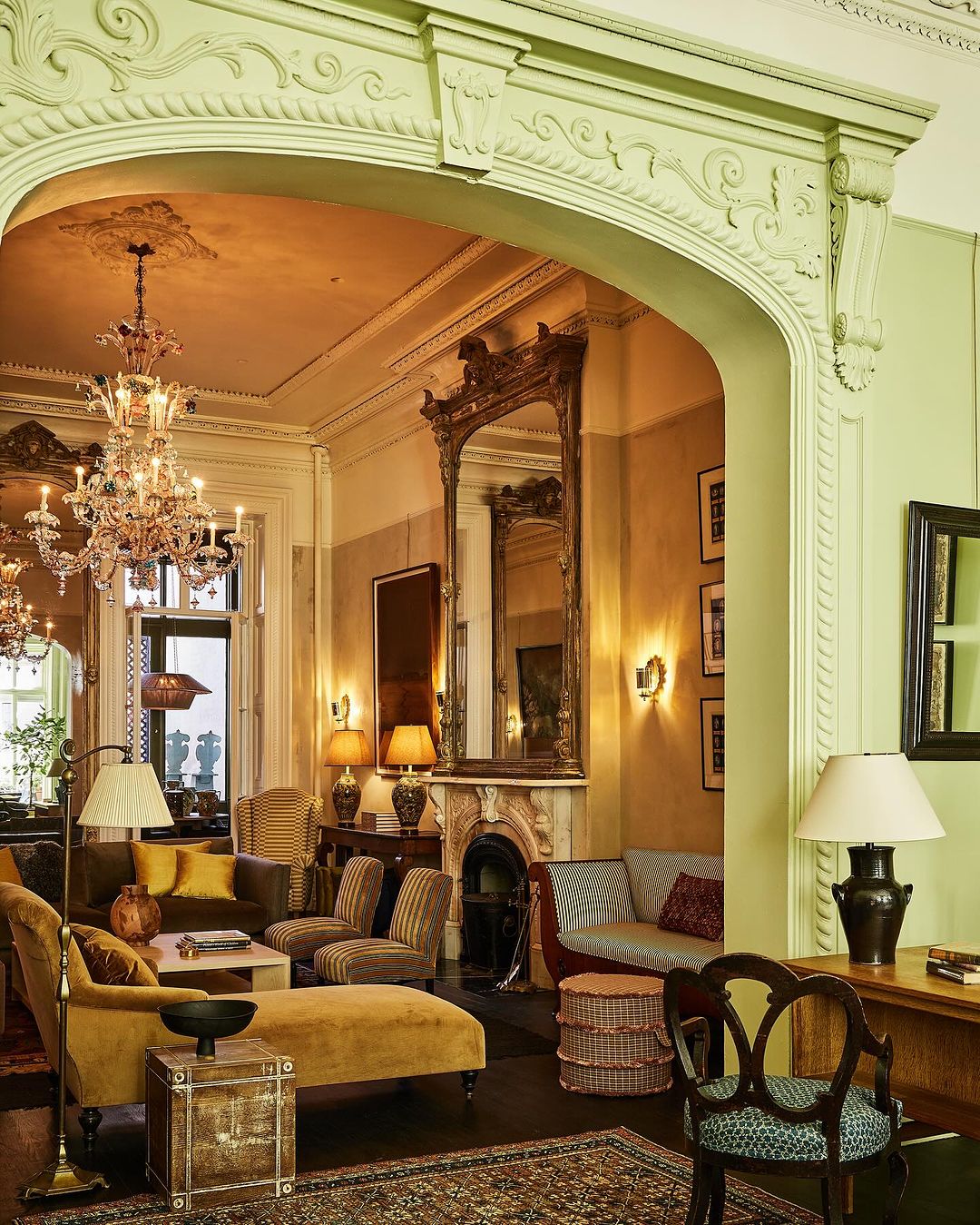 Ornate vintage living room interior with yellow seating, chandeliers, fireplace, large mirrors, carpet, lamps, and decor.