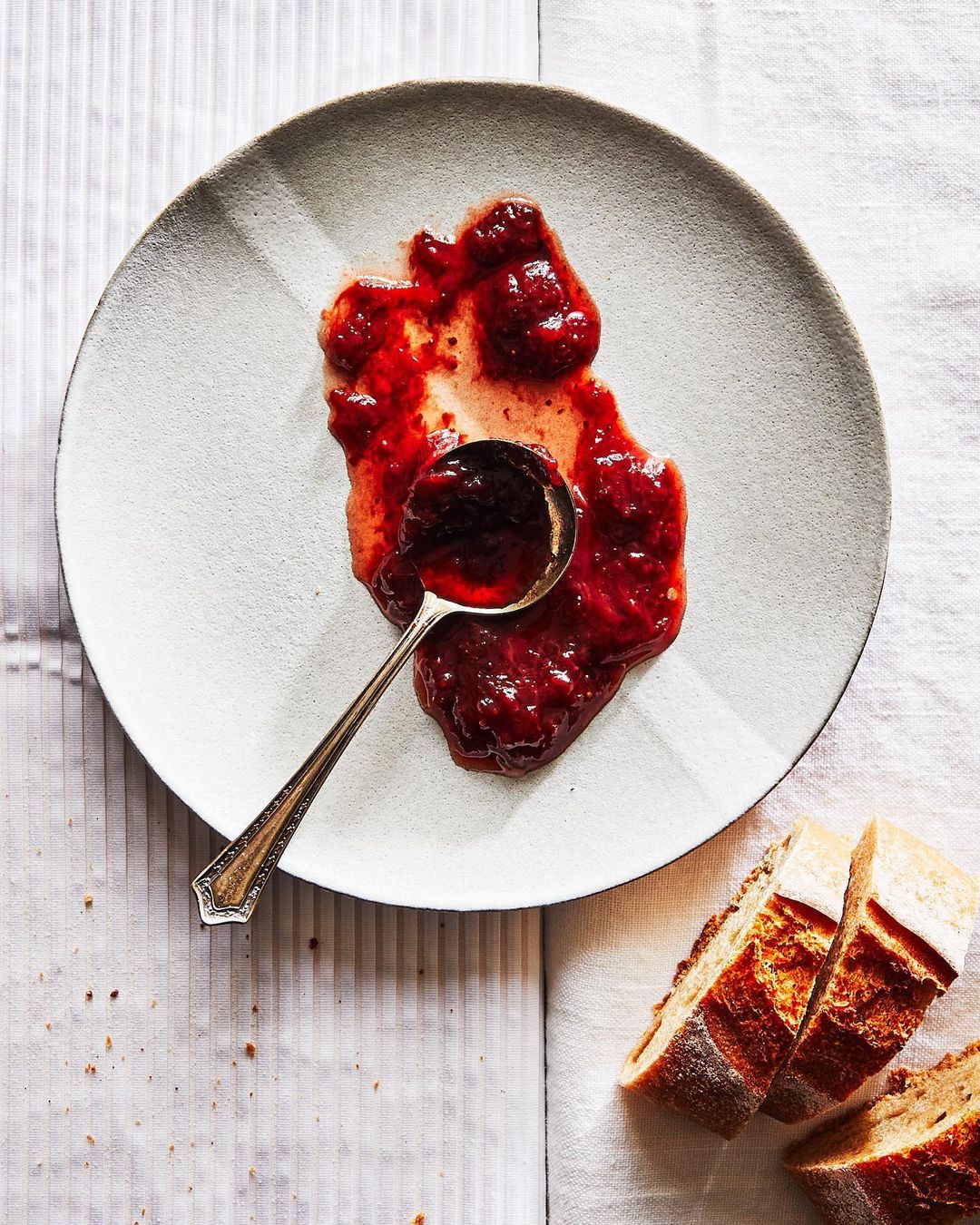 Overhead view of a plate with strawberry jam and a spoon, next to sliced bread on a white linen tablecloth, lit with natural light.