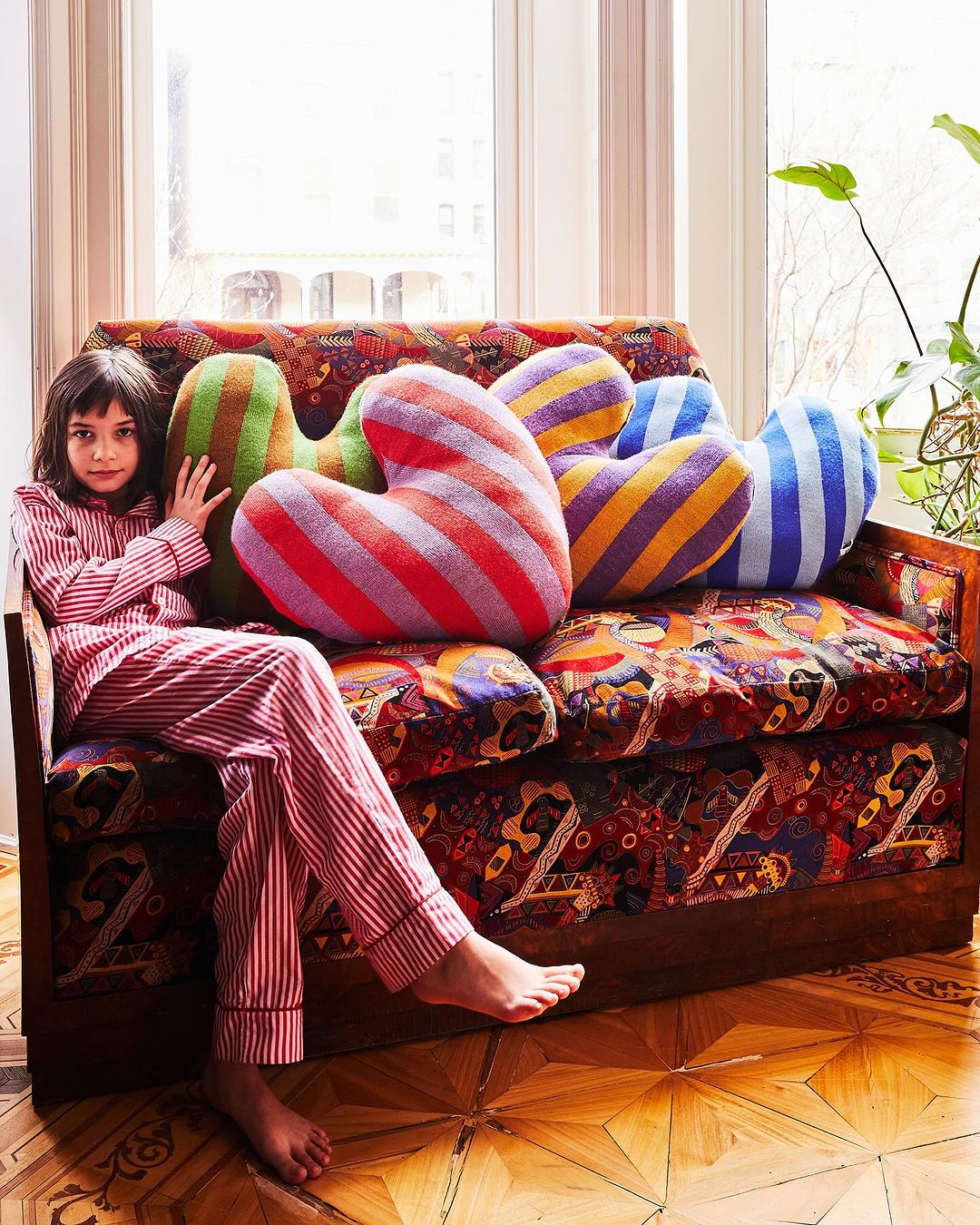 Young girl in red striped pajamas sits on a patterned couch surrounded by colorful striped heart-shaped pillows in a sunlit room.