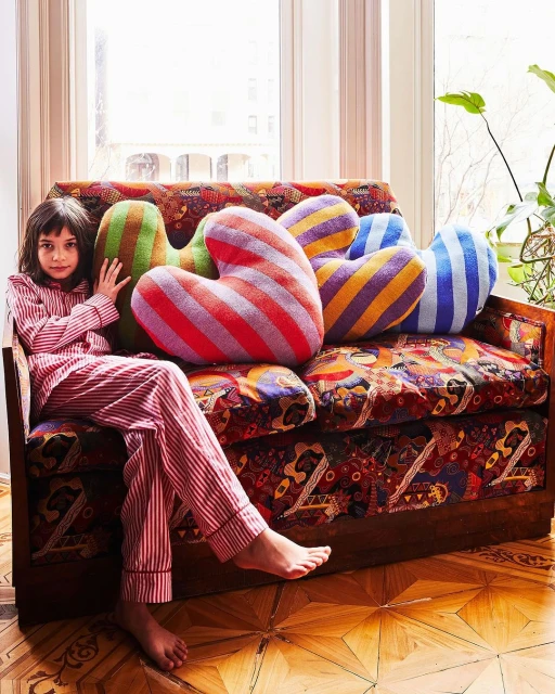 Young girl in red striped pajamas sits on a patterned couch surrounded by colorful striped heart-shaped pillows in a sunlit room.